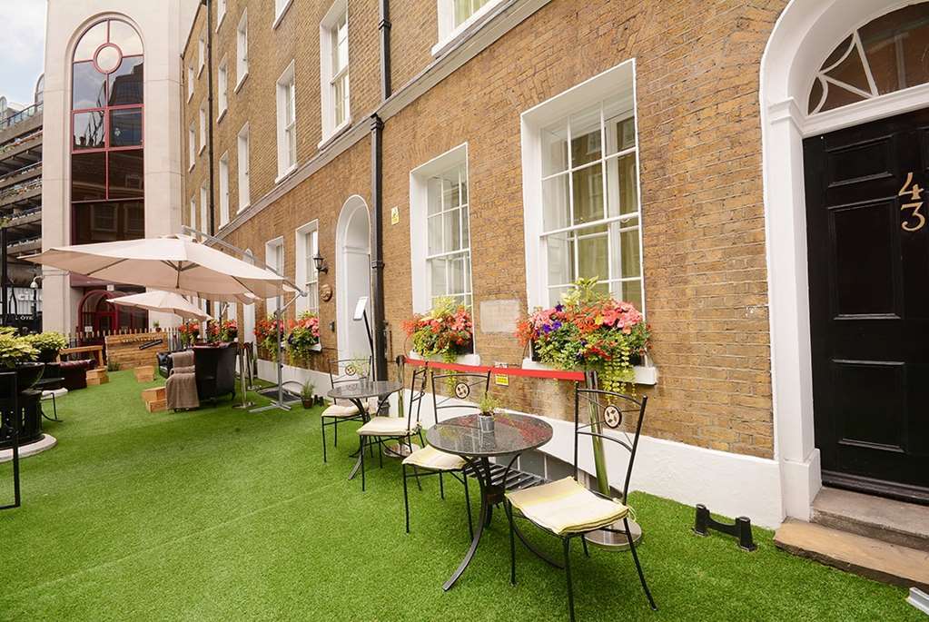 מלון לונדון Montcalm Brewery Townhouse מראה חיצוני תמונה The photo depicts a charming outdoor area in front of a building with a warm, textured brick facade. There is a neatly maintained lawn, likely made of artificial grass, and several small round tables with chairs are arranged outside. Flower pots with