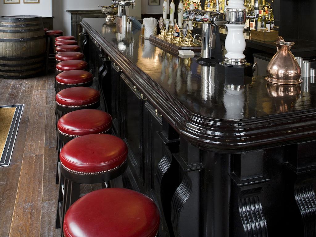 מלון לונדון Montcalm Brewery Townhouse מראה חיצוני תמונה The photo shows a bar setting with a long, polished dark wooden bar counter. Attached to the counter are several bar stools with red leather seats. Behind the bar, there are various bottles, suggesting a well-stocked bar. The flooring appears to be w