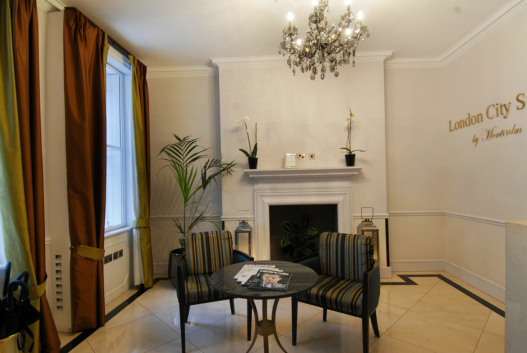 מלון לונדון Montcalm Brewery Townhouse מראה חיצוני תמונה The photo shows a stylish and elegantly designed interior space, likely a lobby or waiting area. It features a chandelier hanging from the ceiling, providing a warm light. There is a white wall with the text "London City by [something]" displayed. 

