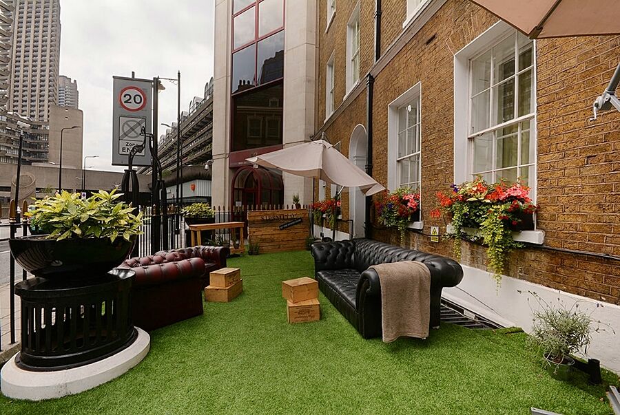 מלון לונדון Montcalm Brewery Townhouse מראה חיצוני תמונה The photo depicts a charming outdoor space, likely a patio or garden area. It features two comfortable-looking leather sofas, one of which is a deep burgundy color, placed on a green artificial lawn. There are wooden crates that could serve as side t